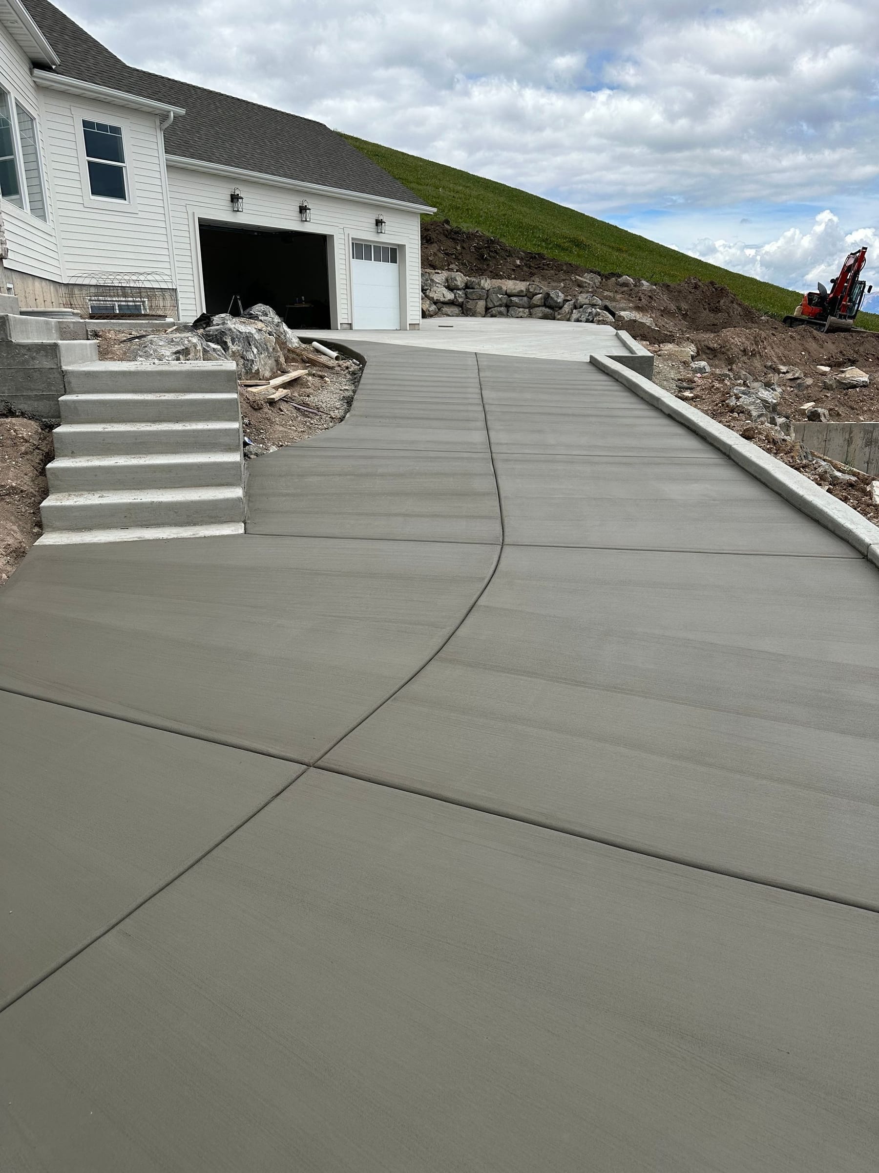 New concrete driveway leading up to a house with a garage and stairs, on a slight slope, under a cloudy sky.