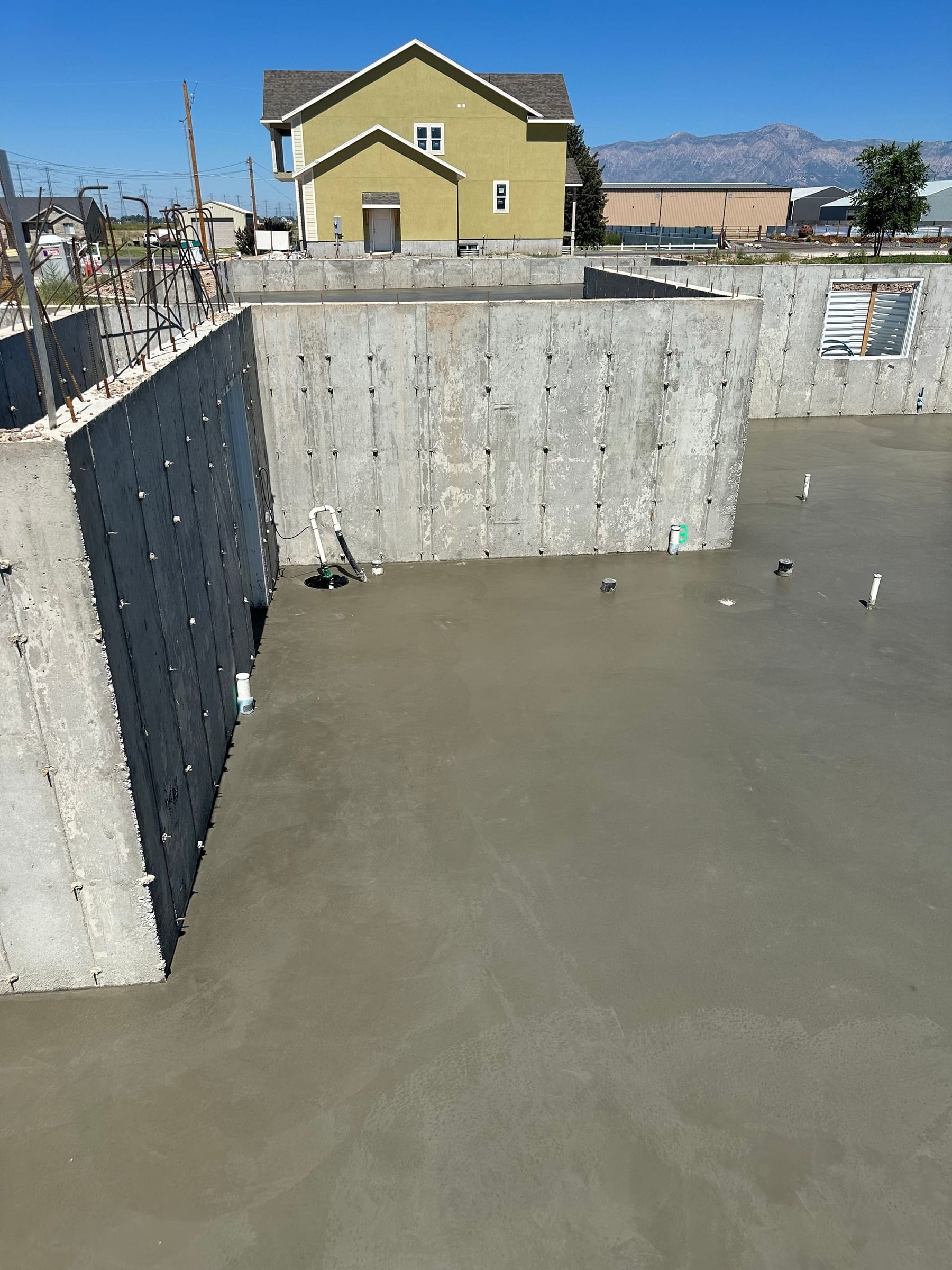 Concrete foundation with a freshly poured floor, walls, and a light yellow house in the background.