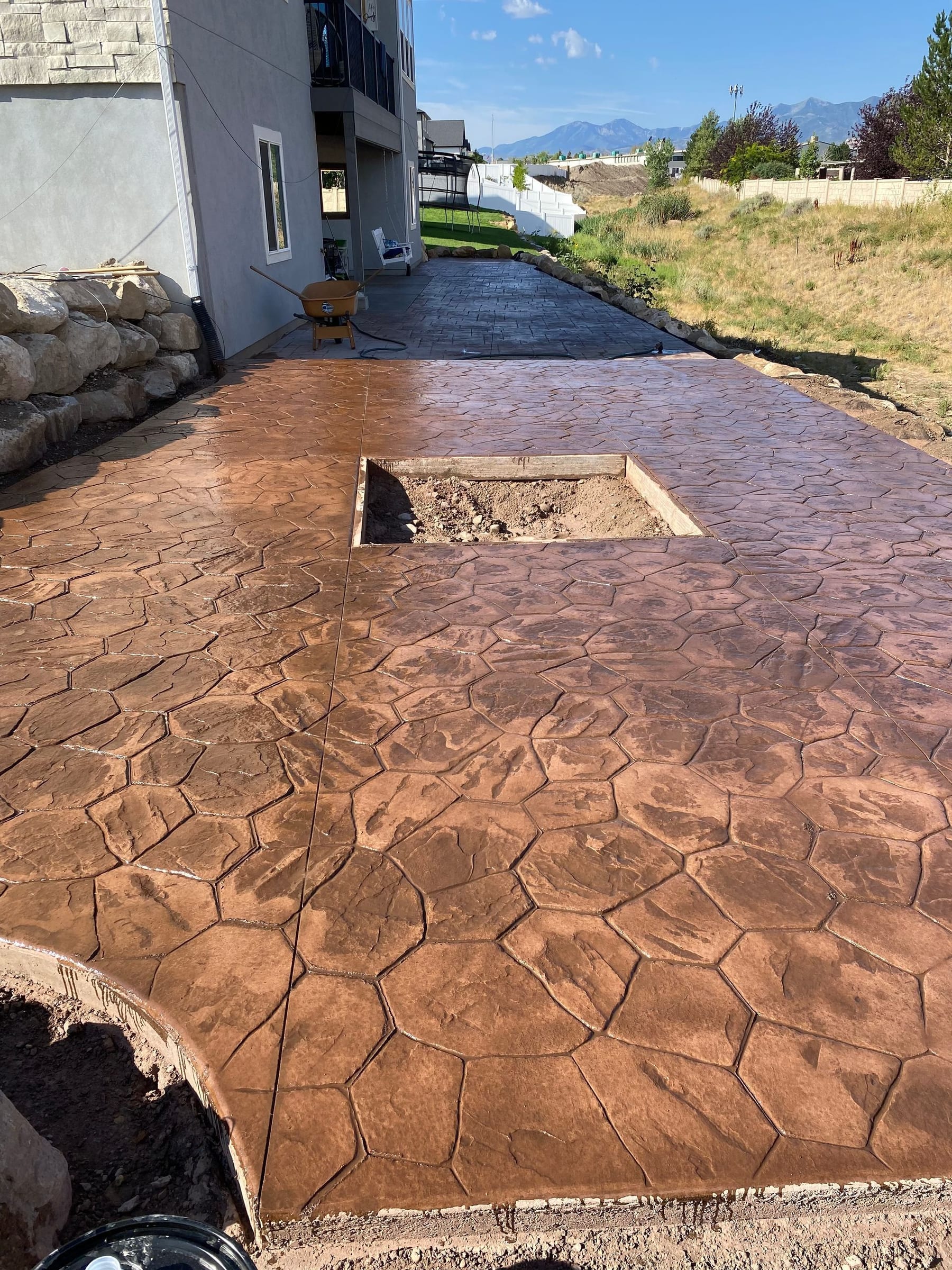 Brown stamped concrete patio with fire pit opening, next to a house with mountain view.